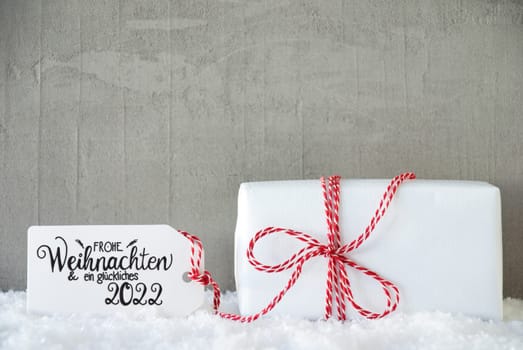 Label With German Calligraphy Frohe Weihnachten Und Ein Glueckliches 2022 Means Merry Christmas And A Happy 2022. One White Gift With Red Bow. Gray Concrete Background With Snow