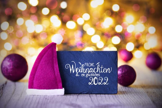 Sighn Wth German Calligraphy Frohe Weihnachten Und Ein Glueckliches 2022 Mean Merry Christmas And Happy 2022. Purple Christmas Ball Ornament With Santa Hat. Sparkling Lights Background
