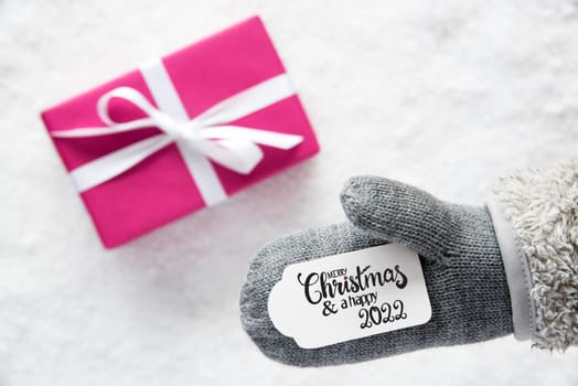 Label With English Calligraphy Merry Christmas And A Happy 2022. Gray Glove With Pink Gift And Snow Background