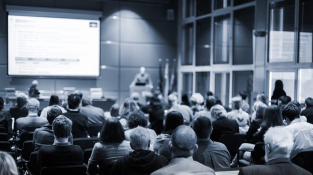 Business and entrepreneurship symposium. Speaker giving a talk at business meeting. Audience in the conference hall. Rear view of unrecognized participant in audience.