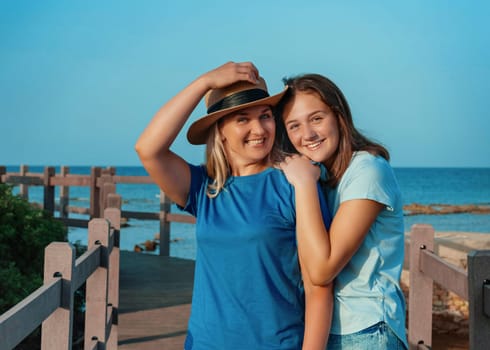Mother wearing straw hat and teenage daughter standing on pier at sea coast, embracing and smiling, t-shirts mockup. Loving family spending time together outdoors in summer, enjoying sunset. Travel concept