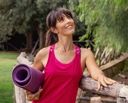 Smiling woman with rolled pilated mat in a park a in a sunny day. Happy middle-aged caucasian women in pink sport clothing outdoor. Healthy lifestyle concept. Mockup