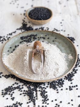 Plate with a scoop of black lentils flour and beans on white wooden table close up. Alternative flour, healthy eating and vegetarian concept. Traditional Asian and North African cuisines ingredient
