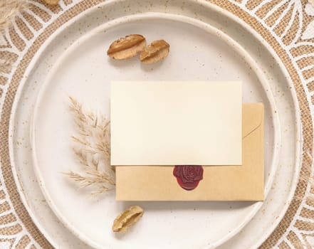 Wedding table place with horizontal blank card and sealed envelope on bohemian placemat top view. Boho wedding card mockup. Tropical beige template with dried pampas grass, place fot text