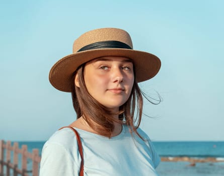 Teen girl standing on wooden walkway by the sea at sunset, wearing light blue t-shirt and straw ha. Summer travel and close to nature concept.