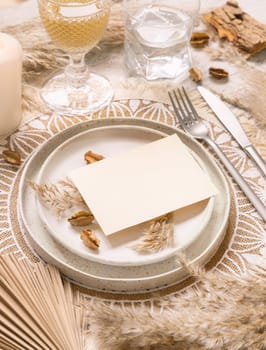 Wedding table place with horizontal blank card on bohemian placemat close up. Boho wedding card mockup. Tropical beige template with dried palm leaves and pampas grass, place fot text