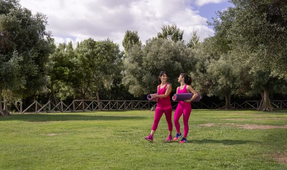 Two women walking in a park a in a sunny day with rolled pilated mats. Young and middle-aged caucasian women in pink clothing outdoor. Sport and healthy lifestyle concept