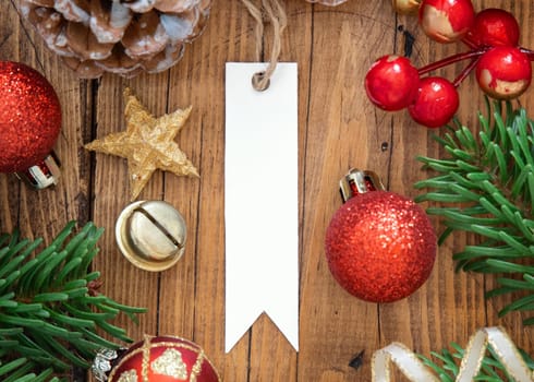 Blank paper gift tag on a brown wooden table with fir tree branches and red and golden Christmas decorations around top view. Winter composition with present label Mockup, copy space