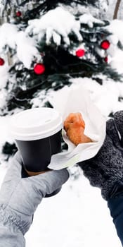 Men's and women's gloved hands hold a cup of coffee and a pie against the backdrop of a Christmas tree in the snow..