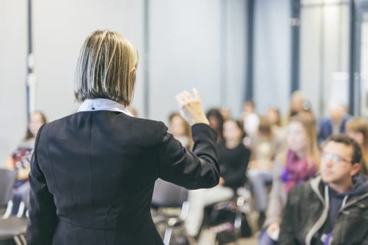 Female speaker giving a talk on corporate business conference. Unrecognizable people in audience at conference hall. Business and Entrepreneurship event.