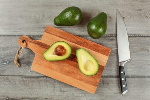 Table top view - avocado cut in half on wooden chopping board, chef knife next to it, two whole green pears above.
