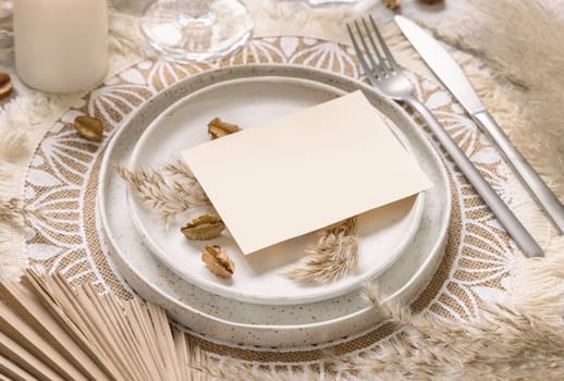Wedding table place with horizontal blank card on bohemian placemat close up. Boho wedding card mockup. Tropical beige template with dried plants, palm leaves and pampas grass, place fot text