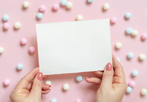 Hands with blank card near pastel pom-poms on pink top view. Romantic scene with blank horizontal card mockup flat lay, place for text . Valentines, Spring or girlish concept