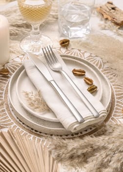 Bohemian Wedding table place with fork and knife over a napkin on bohemian placemat close up. Boho Tropical scene plates, glasses and candle near dried plants, palm leaves and pampas grass