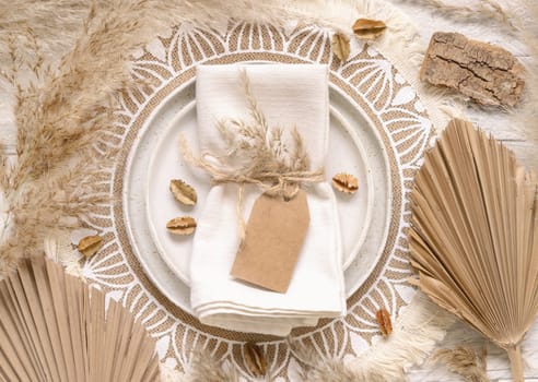 Wedding table place with blank gift tag attached to a napkin on bohemian placemat top view. Boho wedding label mockup Tropical beige template with dried plants, palm leaves and pampas grass, flat lay