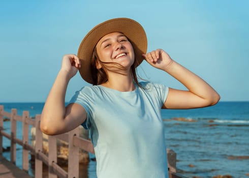 Smiling teen girl standing on wooden pier by the sea at sunset, wearing light blue t-shirt and touching with both hands her straw hat. Summer travel, close to nature concept. T-shirt mockup