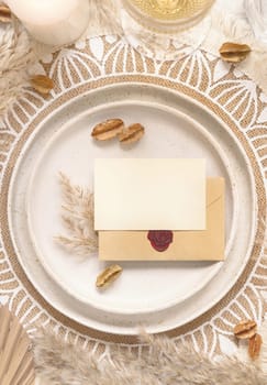 Wedding table place with horizontal blank card and sealed envelope on bohemian placemat top view. Boho wedding card mockup. Tropical beige template with dried plants and pampas grass, place fot text