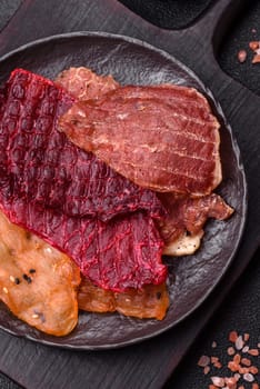 Jerky, dried meat in the form of slices with salt, spices and herbs on a dark concrete background