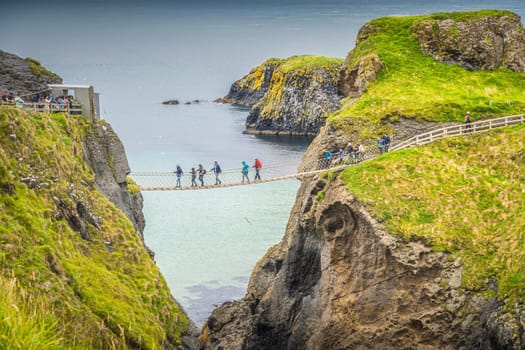 Closeup on people on the Carrick a Rede rope bridge and scenic island surrounded by turquoise Atlantic Ocean, Wild Atlantic Way, Northern Ireland
