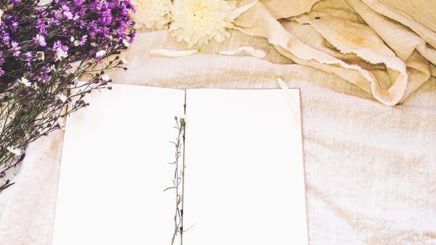 Blank white paper notebook and flower decoration. Greeting card on a white natural  linen background. Top view. Copy space.