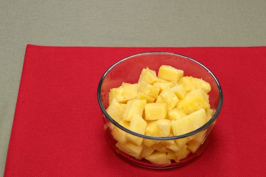One whole sweet pineapple sliced into pieces and place in a clear glass bowl close-up on a red place mat. Sweet yellow pineapple fruit pieces in a round bowl on a red place mat on a green tablecloth.