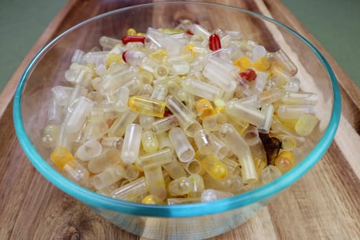 Health supplement capsules and containers accumulated in a clear glass bowl close-up on a table. Emptied with residue health supplement capsules closeup on a wooden serving tray interior in the day.