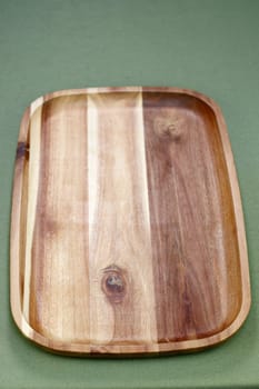 Close-up of a hardwood beige and brown rectangle serving tray with a rail or lip raised edge. Vertical acacia wooden rectangle tray that is used to serve food resting on a green table cloth close-up.