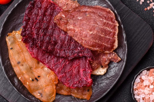 Jerky, dried meat in the form of slices with salt, spices and herbs on a dark concrete background