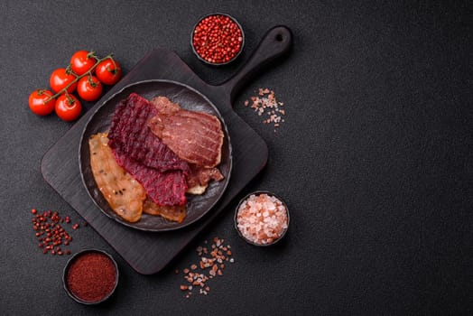 Jerky, dried meat in the form of slices with salt, spices and herbs on a dark concrete background