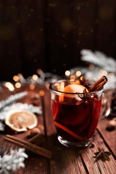 Christmas time concept photo. Glass cup or mug with Mulled Wine inside on wooden table, background. Garlands, Christmas decorations, cinnamon, hazelnut, dried or sublimated orange, spices, white fir