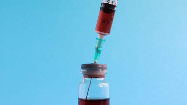A syringe sticks out of a red liquid bottle. Isolated on a blue background. Medicine, injections, vaccines and disposable syringes, drug concept. Sterile bottle. Glass ampoule medical for injection