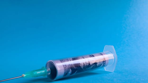 A roll of 100 dollars money inserted into a white medical hypodermic needle on a blue background. The concept of health care costs, credit, investment. Copy space for your text or logo