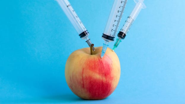 Three different medical syringes sticking deep into an apple on a blue background. The fruit is strewn with syringes. A ripe red apple pierced from all sides by syringes. Healthcare or cosmetology