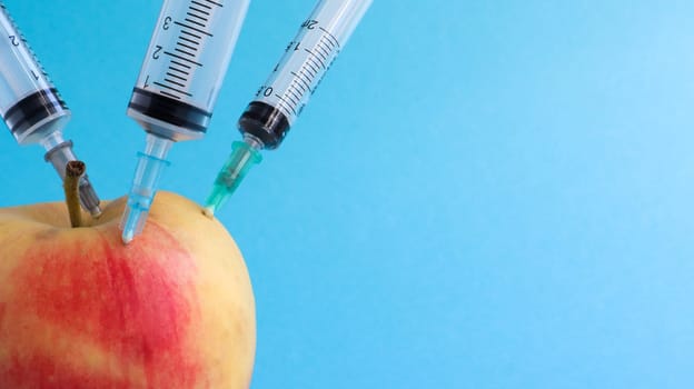 Three different medical syringes sticking deep into an apple on a blue background. The fruit is strewn with syringes. A ripe red apple pierced from all sides by syringes. Healthcare or cosmetology