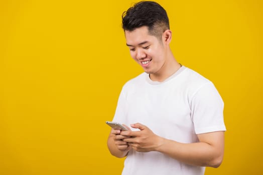 Asian handsome young man smiling positive using smartphone to get in touch screen or typing message, male reading good news message enjoy mobile application, studio shot isolated on yellow background