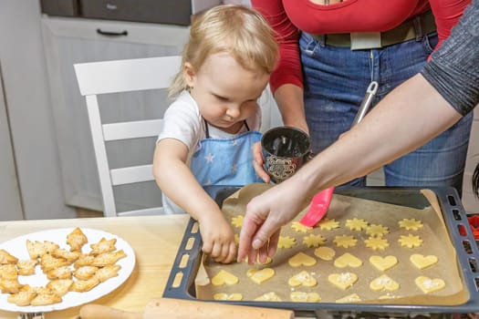 Adorable little child boy puts dough on baking tray with mom and grandmother together in domestic kitchen.