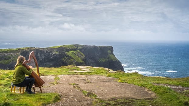 Woman playing harp on the top of iconic Cliffs of Moher, popular tourist attraction, Wild Atlantic Way, County Clare, Ireland