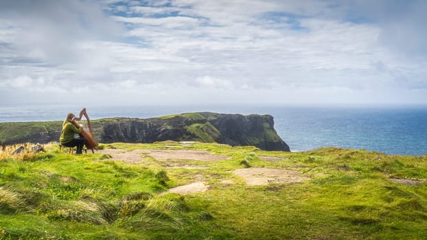 Woman playing harp on the top of the cliff with stunning view on iconic Cliffs of Moher, popular tourist attraction, Wild Atlantic Way, Clare, Ireland