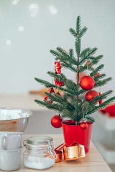 Small decorated Christmas Tree with Christmas toys in red pot placed on kitchen table near cups and sugar. Christmas decoration in the kitchen at home. Concept of celebration of Christmas Holidays.