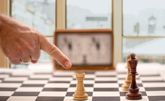 The hand and chessboard with figures and clock on blurred window background. Selective focus