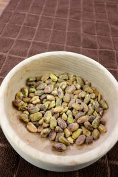 Round wooden bowl of green roasted pistachio nuts without shells closeup on a brown towel on a table. Close-up of shelled roasted pistachio nuts in a wooden bowl resting on a brown towel on a table