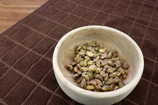 Shelled pistachio nut with sea salt in a round wood bowl on a brown towel close-up. Delicious pistachios and sea salt without shells closeup in a round wooden bowl on a brown towel resting on a table.