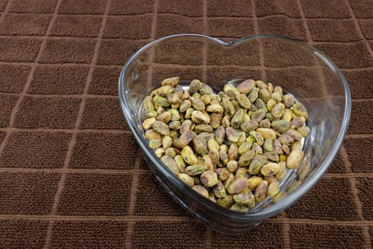 Roasted pistachios without shells with sea salt in a clear glass heart shaped dish on a brown cloth close-up. Nutritious shell-less roasted pistachios with sea salt in a heart shaped clear glass dish