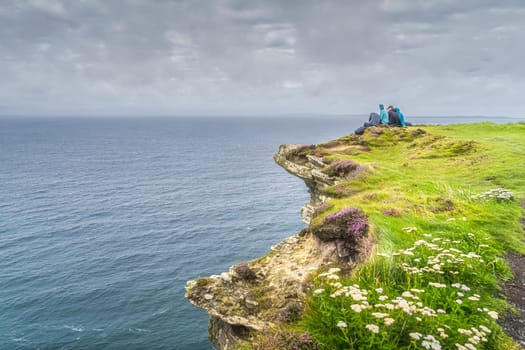 Group of young people sitting on the edge of cliff on iconic Cliffs of Moher, popular tourist attraction, Wild Atlantic Way, County Clare, Ireland