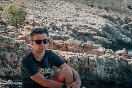 A man sits and hugs his knee against the backdrop of the rocky mountains.