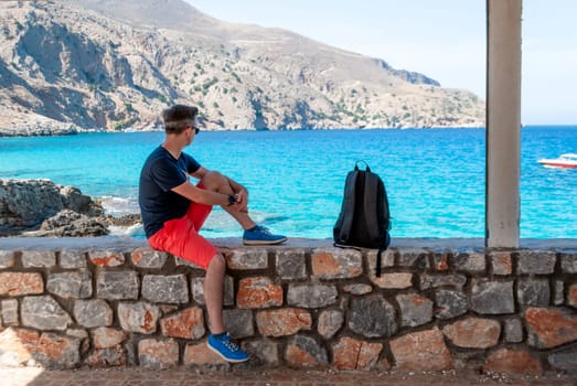 A man in red shorts sits on a stone fence and looks out over the turquoise sea