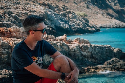 A man in sunglasses against the backdrop of rocks and the turquoise sea.