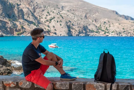 A man looks at the turquoise sea and mountains sitting on a stone parapet.