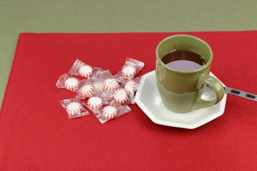 Hot cocoa beverage of a chocolate holiday mix served in a green coffee cup near peppermint candy on a red placemat on a green tablecloth. Hot chocolate cocoa mix beverage with peppermint candy rounds..
