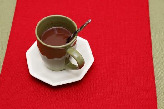 A cup of hot cocoa mix with spoon on a white saucer placed on a red place mat on a green tablecloth. Warm hot chocolate mix in a cup with a metal spoon on a red and green textile tabletop background.